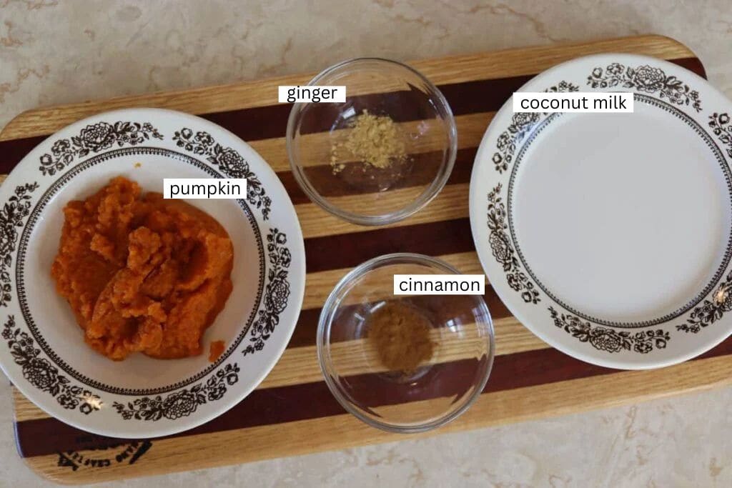 Four plates and bowls on a wooden board hold mashed pumpkin, ground cinnamon, ground ginger, and coconut milk. Each ingredient is labeled with text above or beside it. The surface underneath is light-colored.