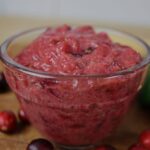 Deep pink sauce with red chunks in it in a glass condiment dish with cranberries spread around it.