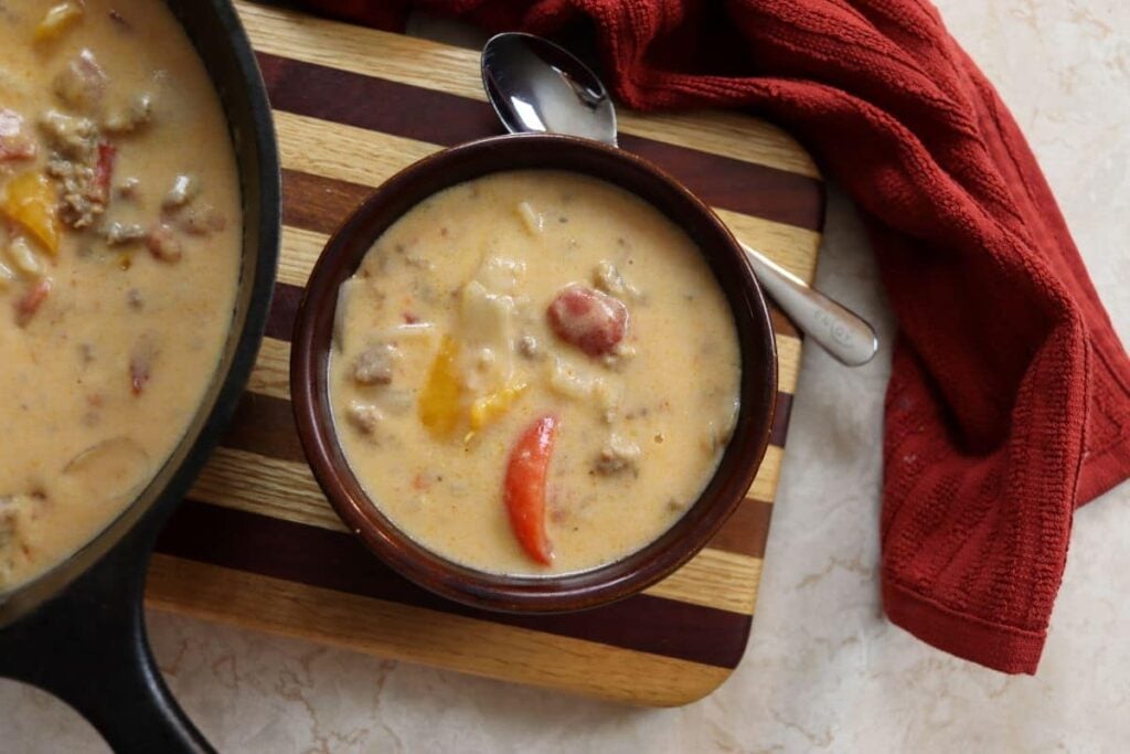 A bowl of creamy soup with chunks of meat, tomatoes, and bell peppers sits on a striped wooden board beside a spoon and a red cloth, with a cast iron skillet partially visible on the side.