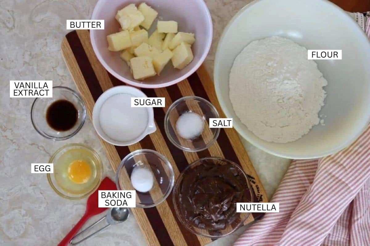 Labeled baking ingredients: butter (cubed), flour, sugar, salt, vanilla extract, egg, baking soda, and Nutella, all arranged on a wooden board and a marble countertop.