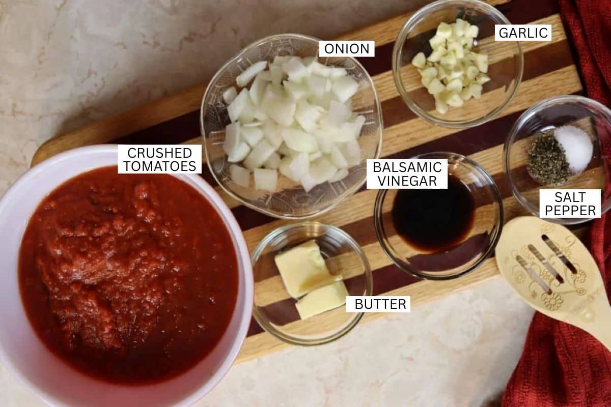 A wooden board holds bowls containing crushed tomatoes, chopped onion, minced garlic, butter, balsamic vinegar, and a mix of salt and pepper, each labeled with text. A red cloth is partially visible on the right.
