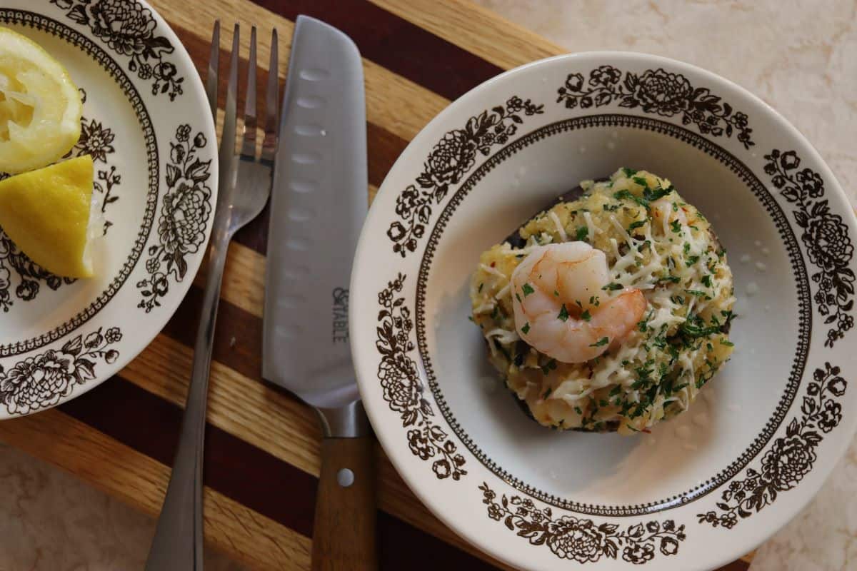 A bowl with a stuffed mushroom topped with shrimp and herbs sits on a wooden board next to a knife, fork, and a plate with lemon wedges.