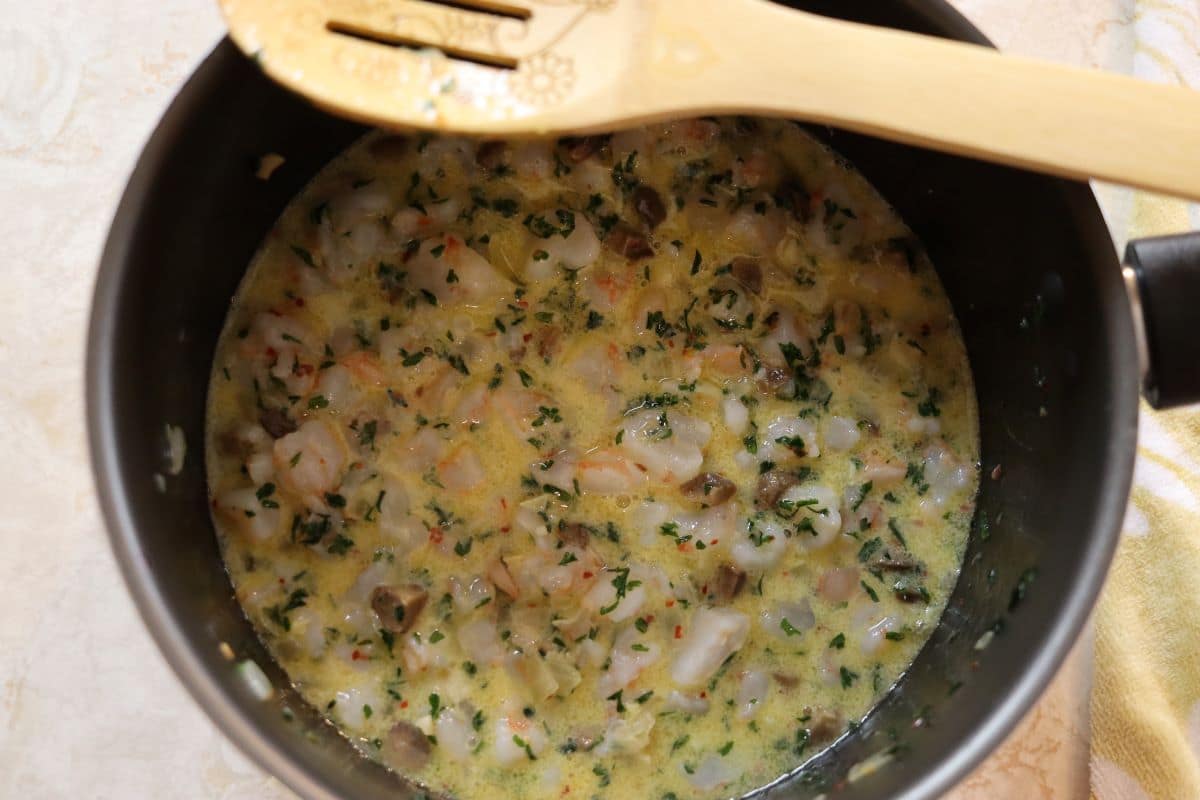Buttery sauce with cut up shrimp and fresh parsley visible in it in a pan with a wooden spoon resting on the side.