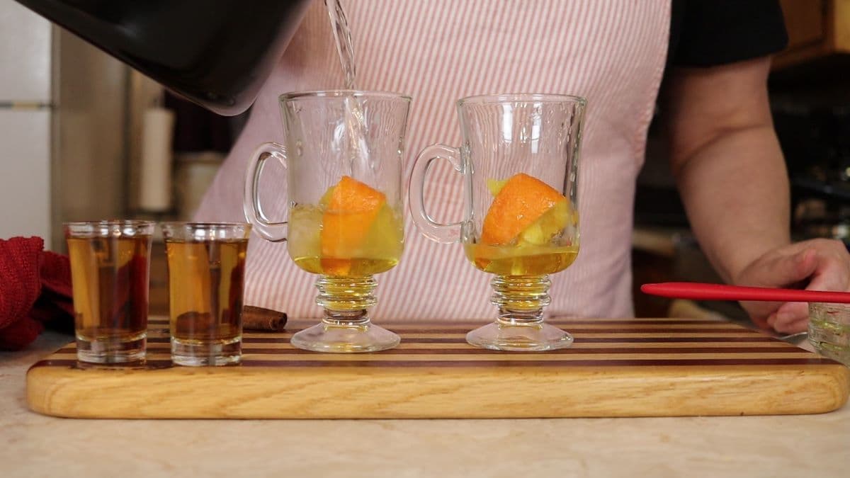 woman pouring hot water into glass cups with honey and oranges in them.