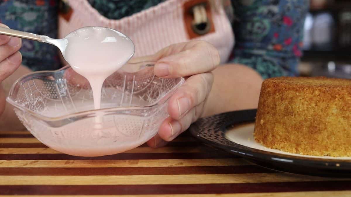 Woman dripping pink icing from a spoon into a small bowl over a striped cutting board with a small cake visible next to it.