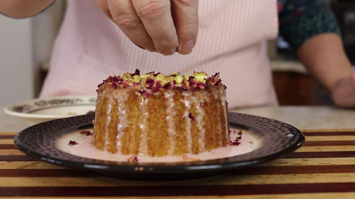 Woman garnishing a small cake that has pink icing dripping down it's sides with red rose petals and crushed pistachios.