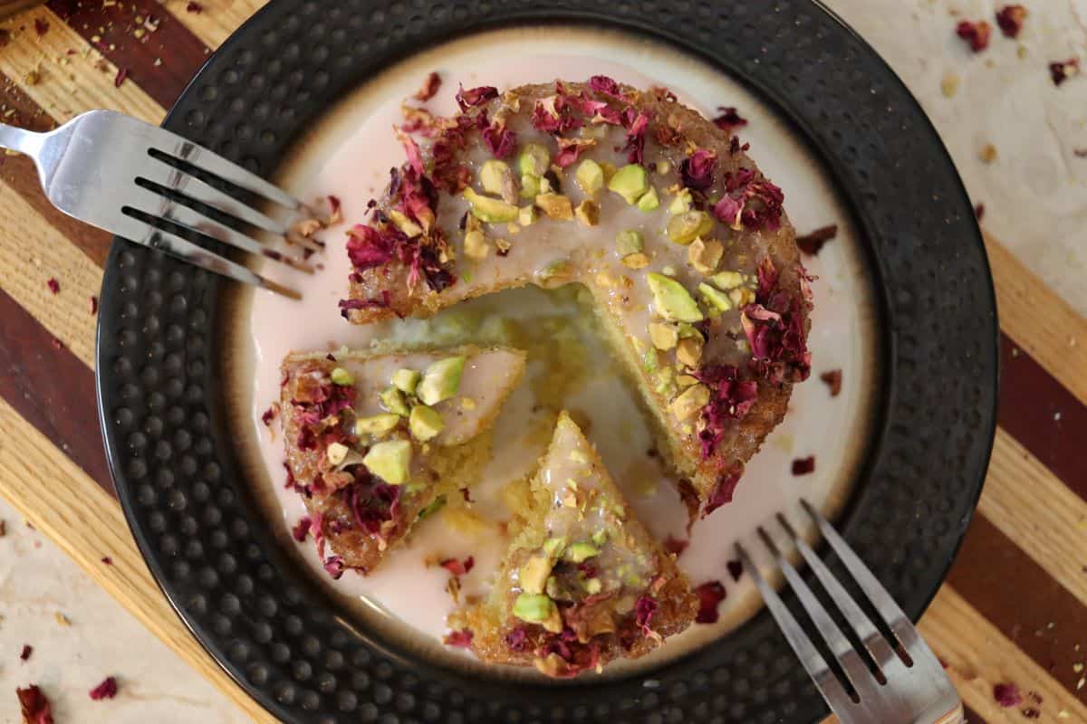 A round dessert topped with chopped pistachios and dried rose petals sits on a black-rimmed plate. A knife and fork rest on a striped wooden surface beside the plate.