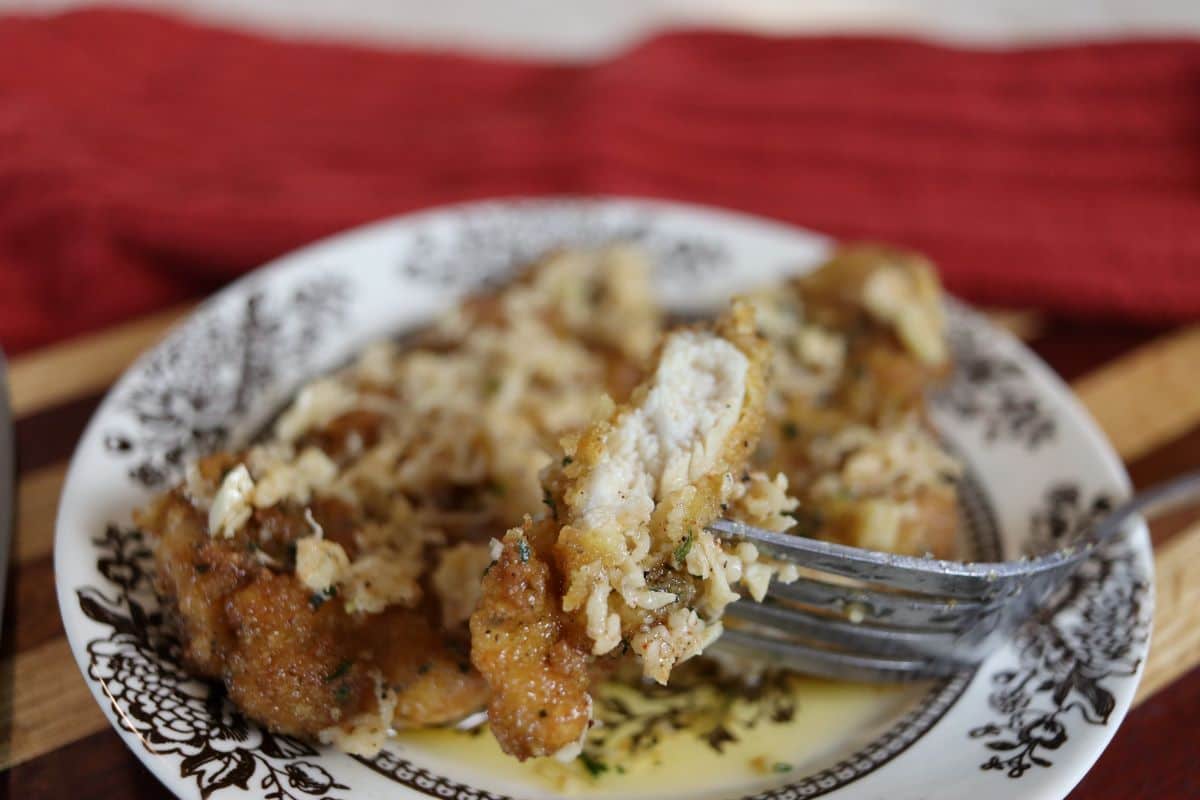 A piece of chicken with parmesan cheese on it on a fork with a plate of chicken in butter sauce and parmesan cheese in the background.