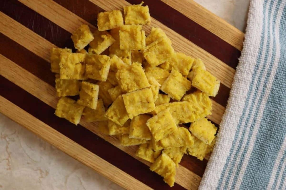 A pile of square, orange colored homemade cat treats on a striped wooden cutting board with a blue and white striped towel next to them.