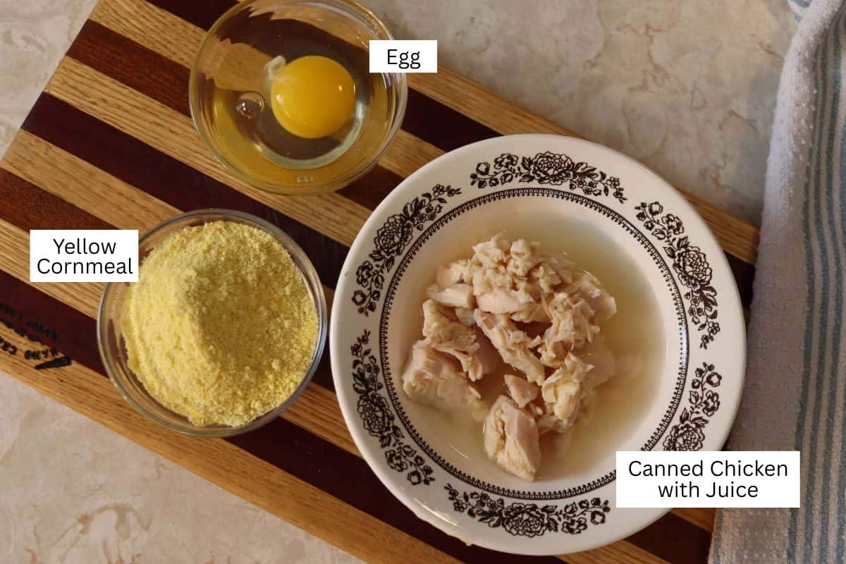 A bowl of canned chicken with juice, a bowl of yellow cornmeal, and a bowl with a raw egg, all labeled, are arranged on a wooden board with a striped towel nearby.