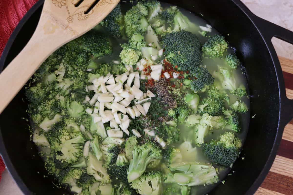 Broccoli, garlic, and spices in a cast iron dutch oven sitting on a striped cutting board with a spoon resting on the side.