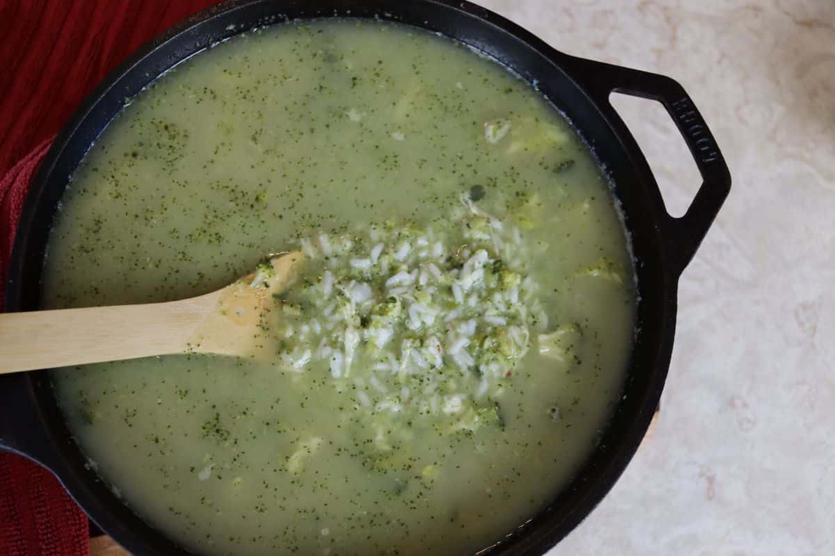 A cast iron pot filled with green soup containing herbs, rice, and vegetables, with a wooden spoon stirring the mixture. The pot is placed on a light-colored surface.