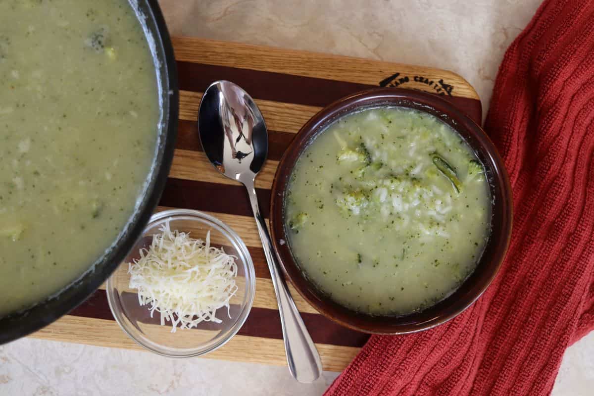 A bowl of broccoli soup on a wooden board, with a spoon, shredded cheese in a small dish, and a red cloth beside it. A pot of soup is partially visible on the left.