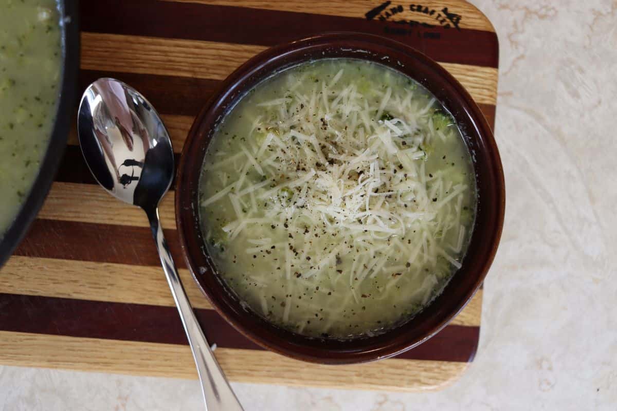 Brown bowl with broth and shredded cheese in it sitting on a striped cutting board with a spoon next to it.