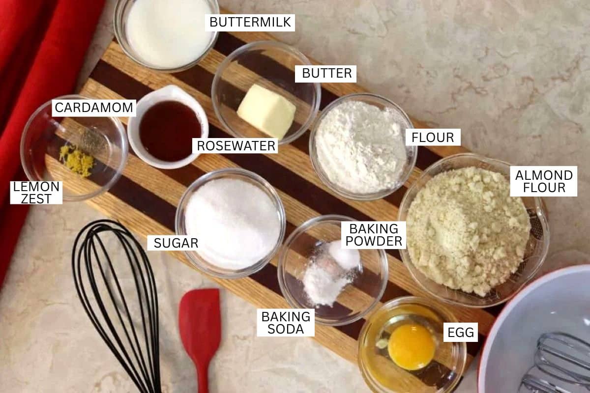 A wooden board holds labeled bowls of ingredients: buttermilk, butter, flour, almond flour, egg, baking soda, sugar, lemon zest, cardamom, rosewater, and baking powder, with a whisk and spatula nearby.