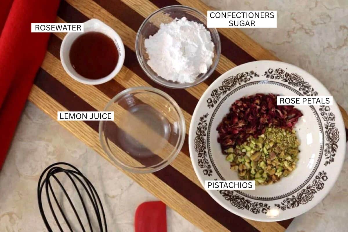A striped wooden tray holding labeled ingredients: rosewater in a small white bowl, confectioners sugar in a glass bowl, lemon juice in a glass, and a plate with rose petals and chopped pistachios.