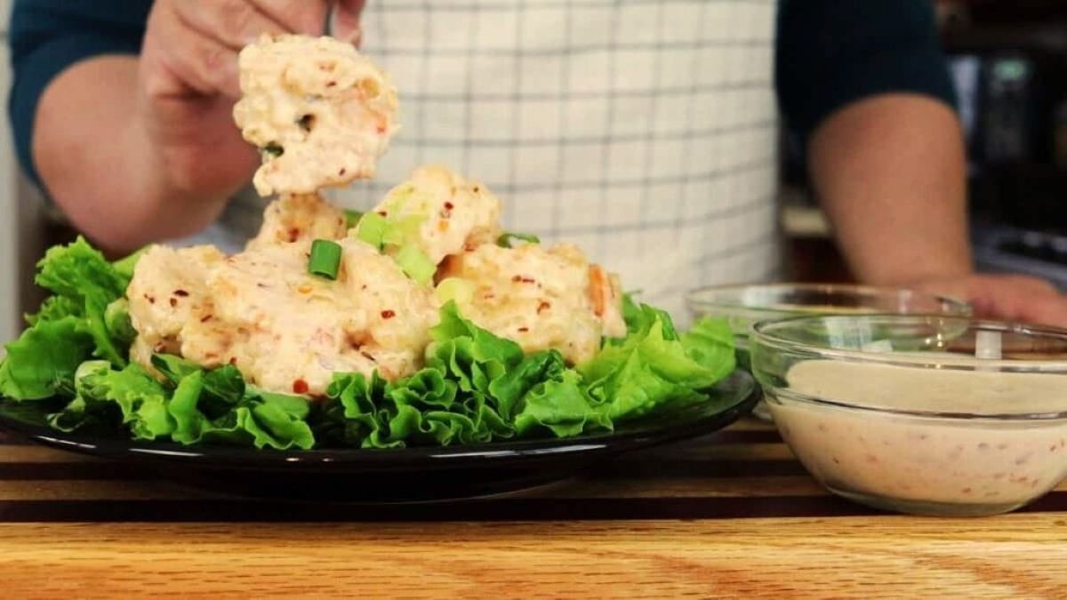 A person in an apron serves creamy, sauced shrimp on a bed of lettuce leaves, with bowls of dipping sauce on the side, all arranged on a wooden countertop.