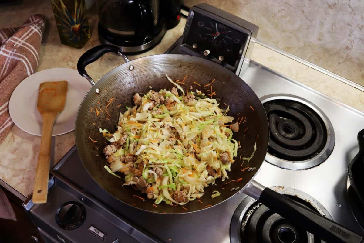 Wok on a stove with ground pork, cabbage, and shredded carrots cooking in it.