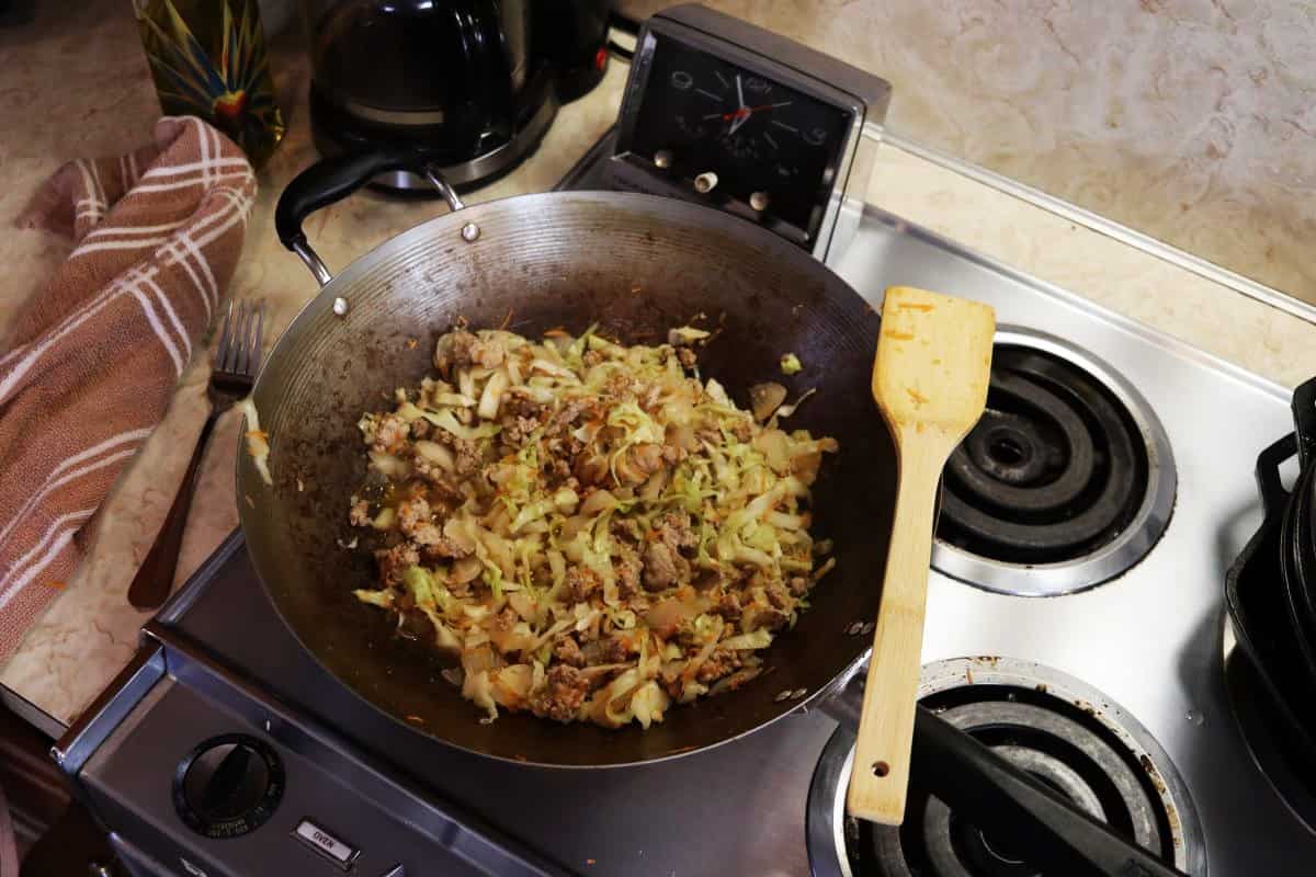 Wok on a stovetop with cooked pork, cabbage, and carrots in it and a wooden spoon resting on the side of it.