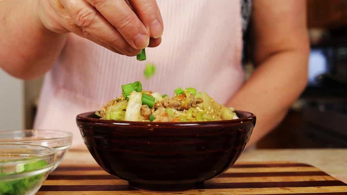 Woman sprinkling chopped scallions over a bowl of pork, cabbage, shredded carrots, and sesame seeds.