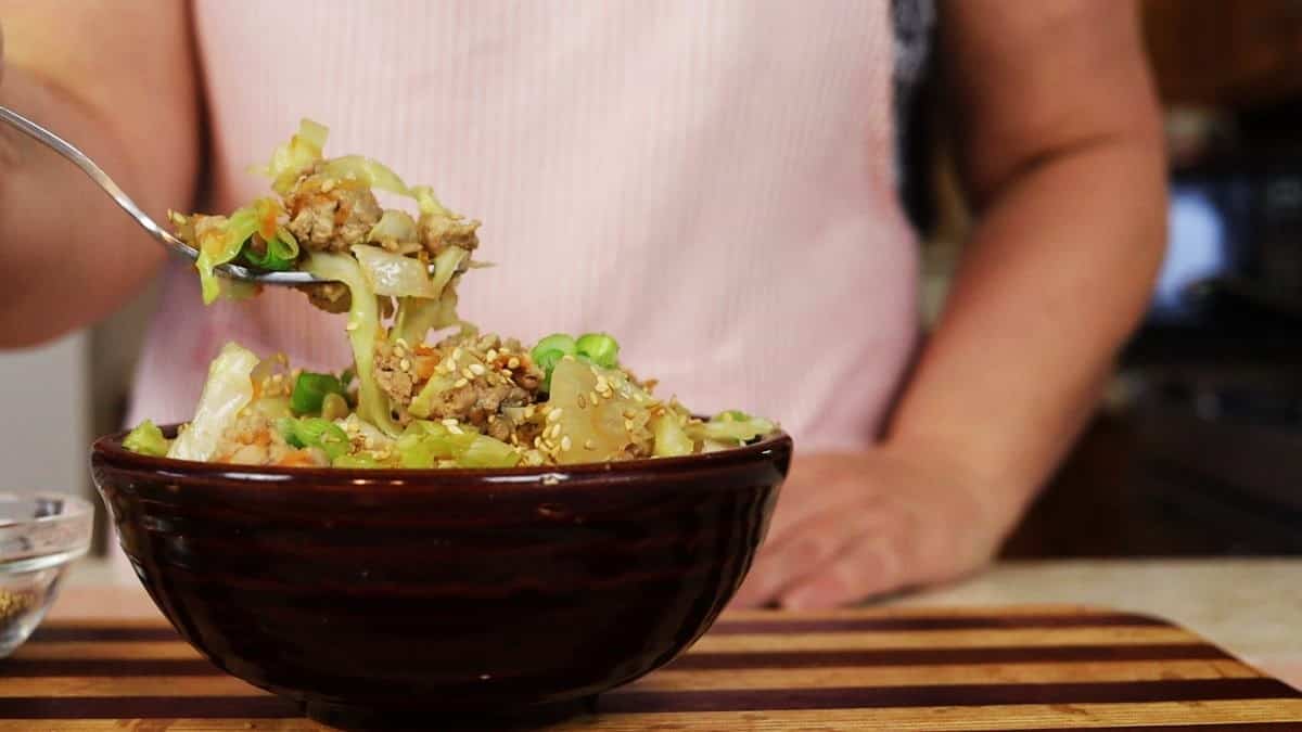 A person holding a spoonful of a vegetable and meat dish topped with green onions and sesame seeds over a dark bowl on a striped wooden surface.