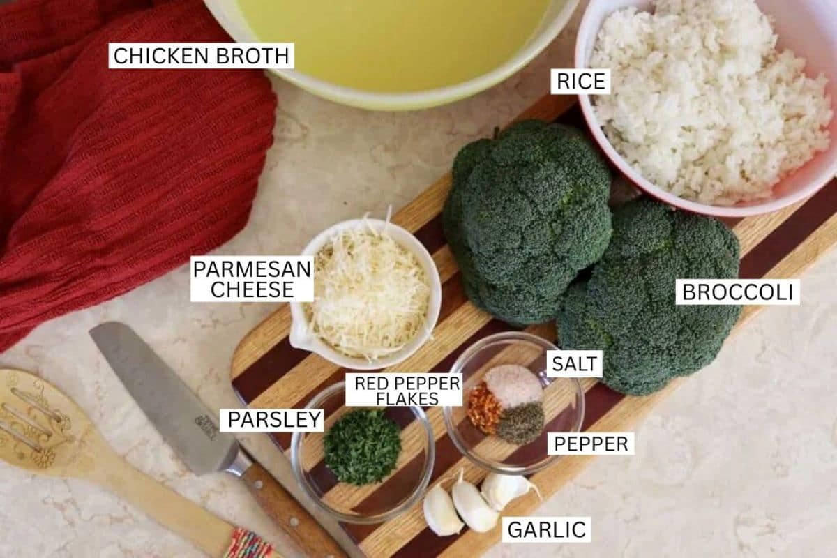 A wooden board holds bowls of cooked rice, chicken broth, broccoli, parmesan cheese, parsley, red pepper flakes, salt, pepper, and garlic. A knife, spoon, and red towel are also visible. Each ingredient is labeled.