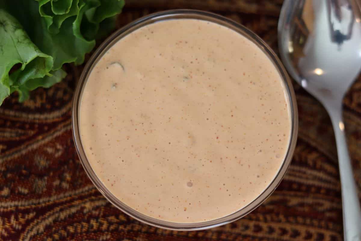 A glass bowl filled with creamy, light pink sauce sits on a patterned surface next to a lettuce leaf and a silver spoon.