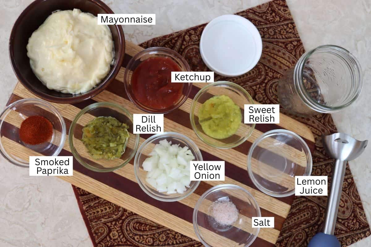A cutting board with labeled bowls containing mayonnaise, ketchup, dill relish, sweet relish, smoked paprika, chopped yellow onion, lemon juice, and salt, along with a stick blender.