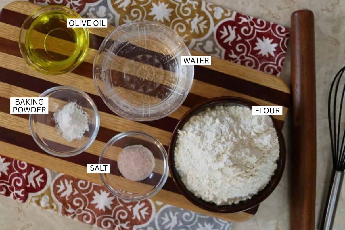 Striped wooden board holding bowls of flour, water, olive oil, baking powder, and salt, all labeled. A whisk is nearby on a patterned cloth.