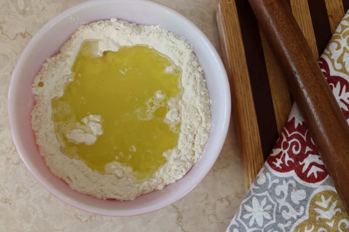 Flour with a well full of oil and water in it in a white bowl sitting next to a striped cutting board and a red and grey cloth.