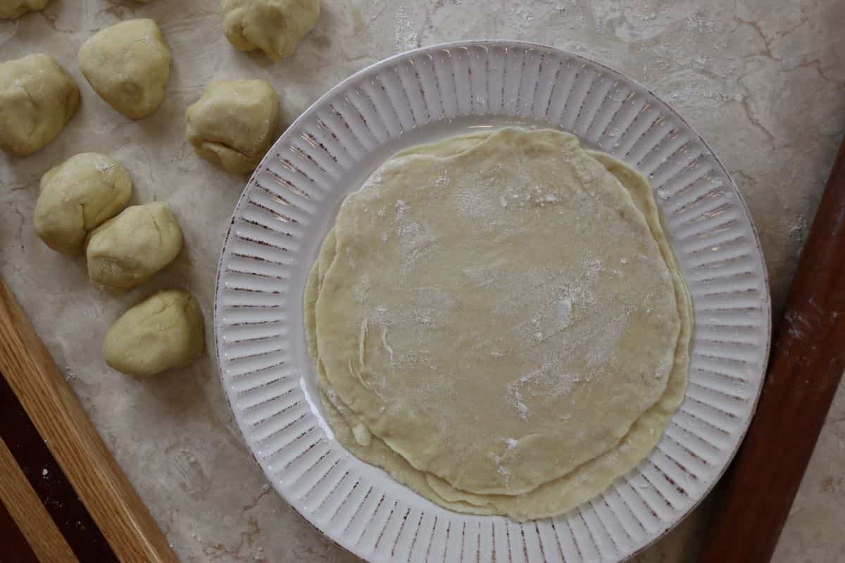 Raw tortilla shells stacked on a white plate with flour on them. There are dough balls sitting to the side of the plate.