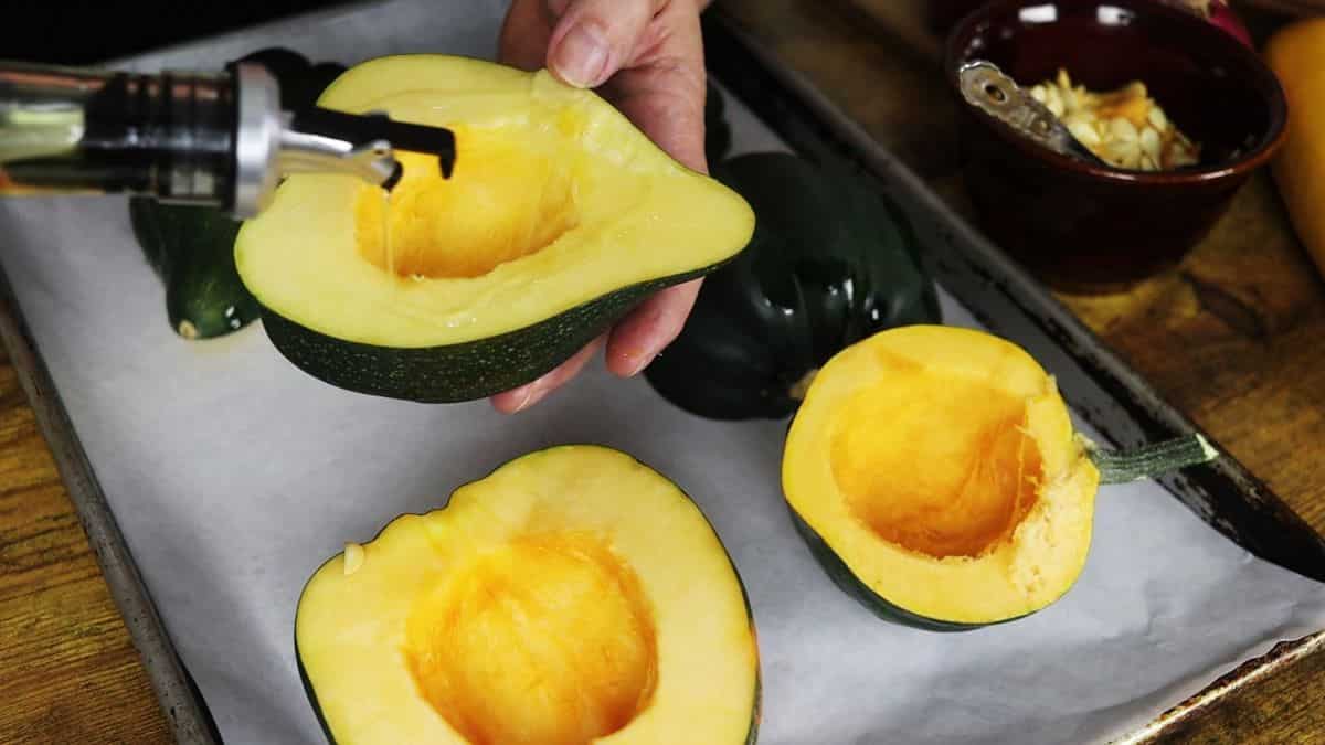 Hands drizzling oil over the flesh of an acorn squash that has been cut in half. Other squash sit under it on a parchment lined baking sheet.
