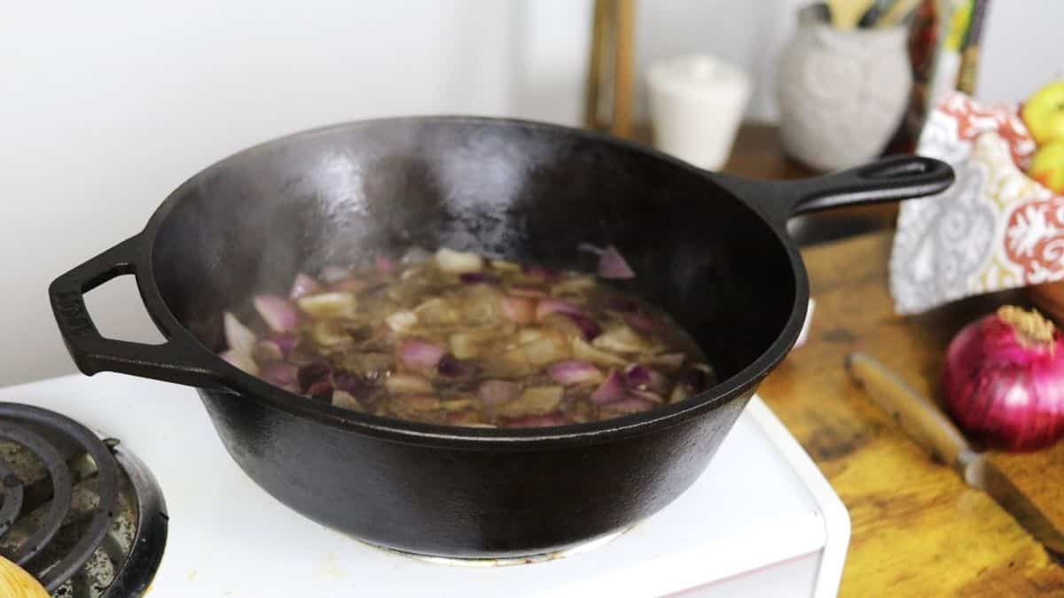 Chopped red onions simmering in liquid in a cast iron pot on a stovetop.