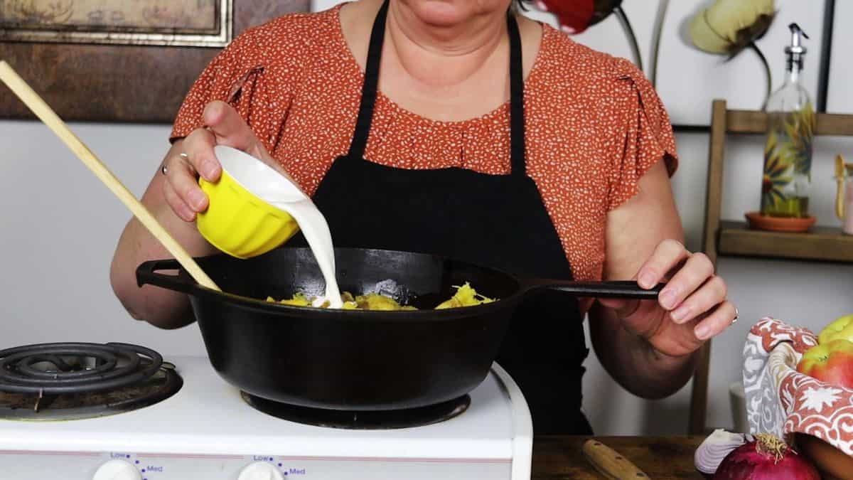 Woman pouring cream into a cast iron pot of acorn squash on a stovetop.