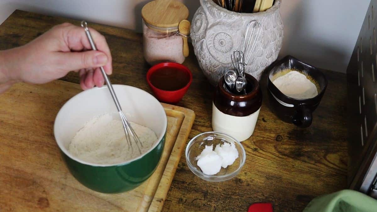 Flour being whisked in a bowl on a counter with small dish of lard, small red bowl, crock of utensils, and a glass canister of pink salt nearby.