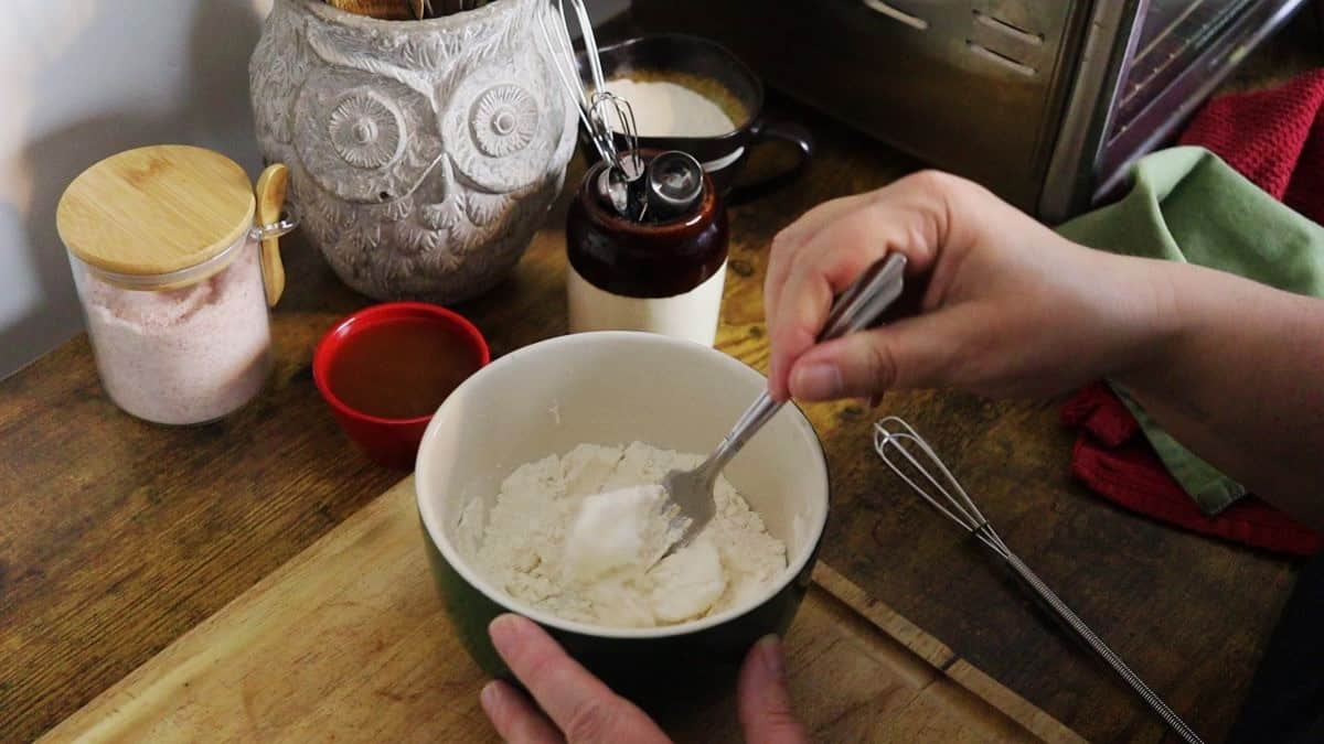 Hand mixing lard into flour in a small bowl with a fork with kitchen utensils in the background.