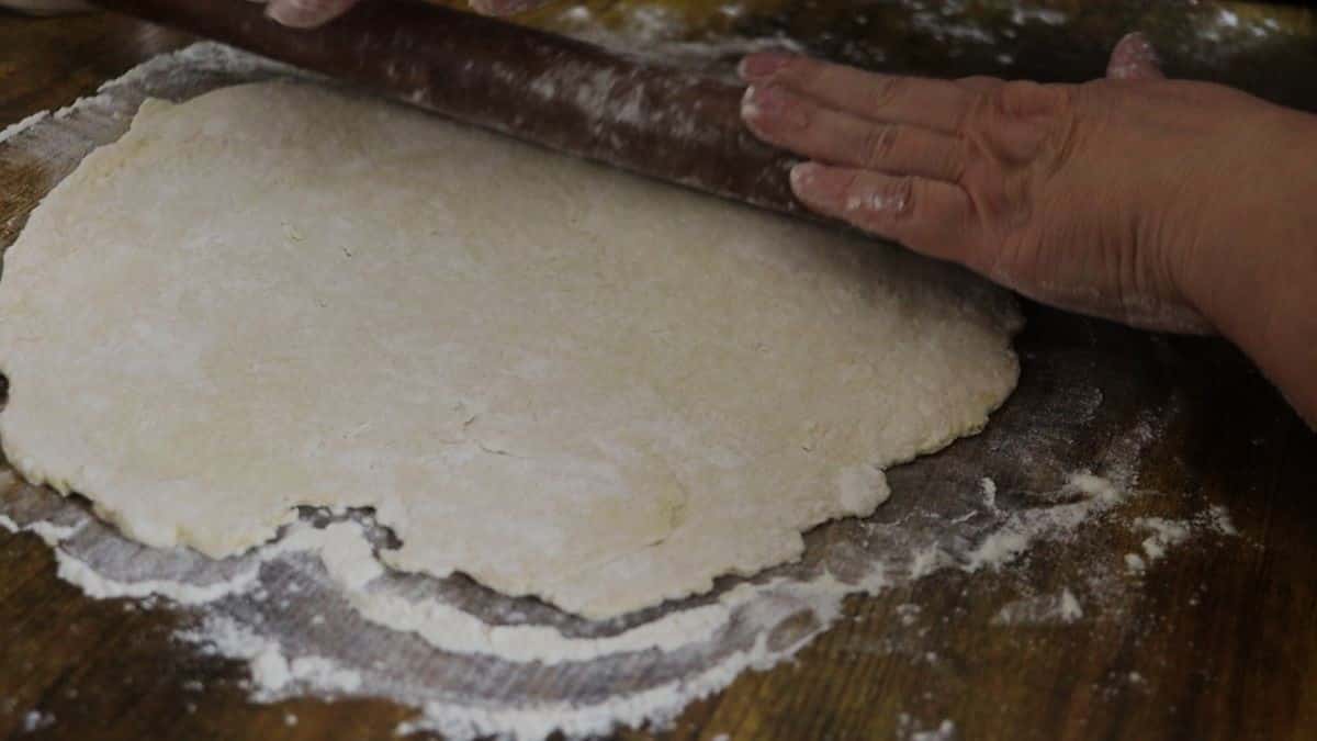 Hands using a rolling pin to roll out dough on a flour covered wooden surface.