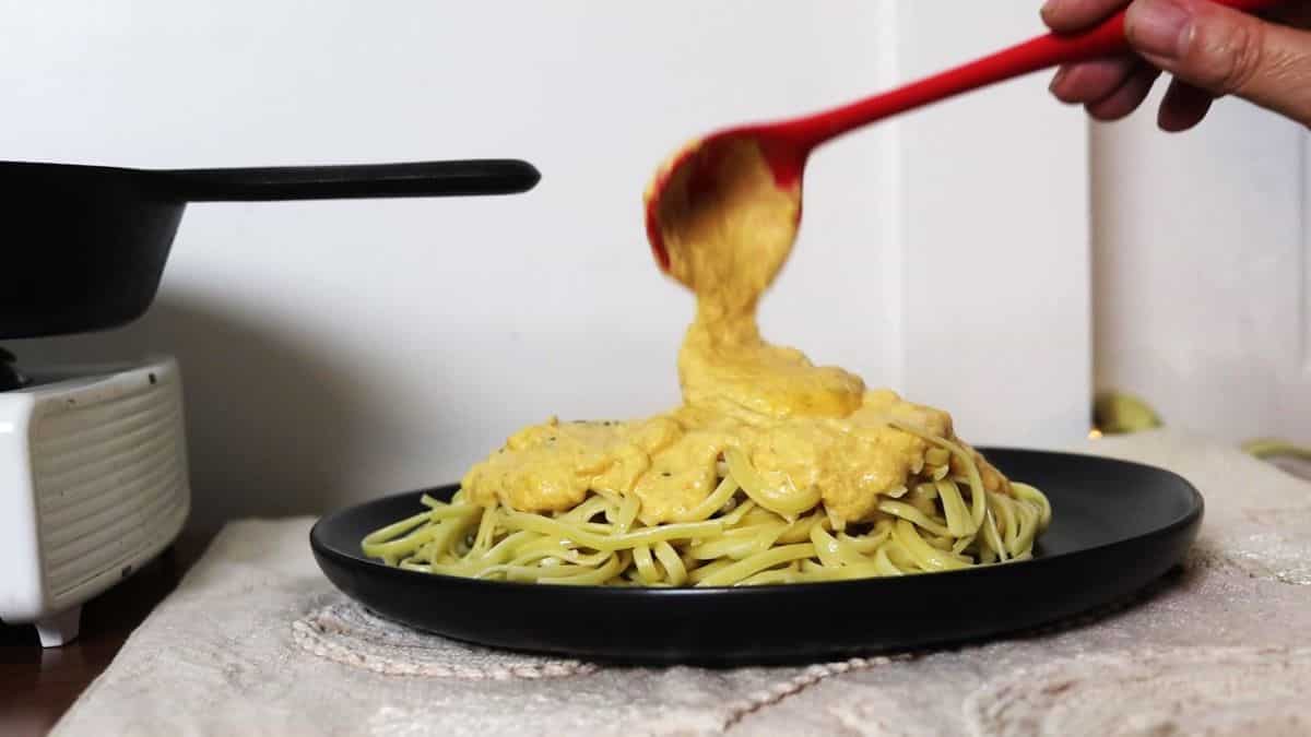 A hand uses a red ladle to pour creamy orange sauce over linguine pasta, set on a black plate atop a light-colored cloth.