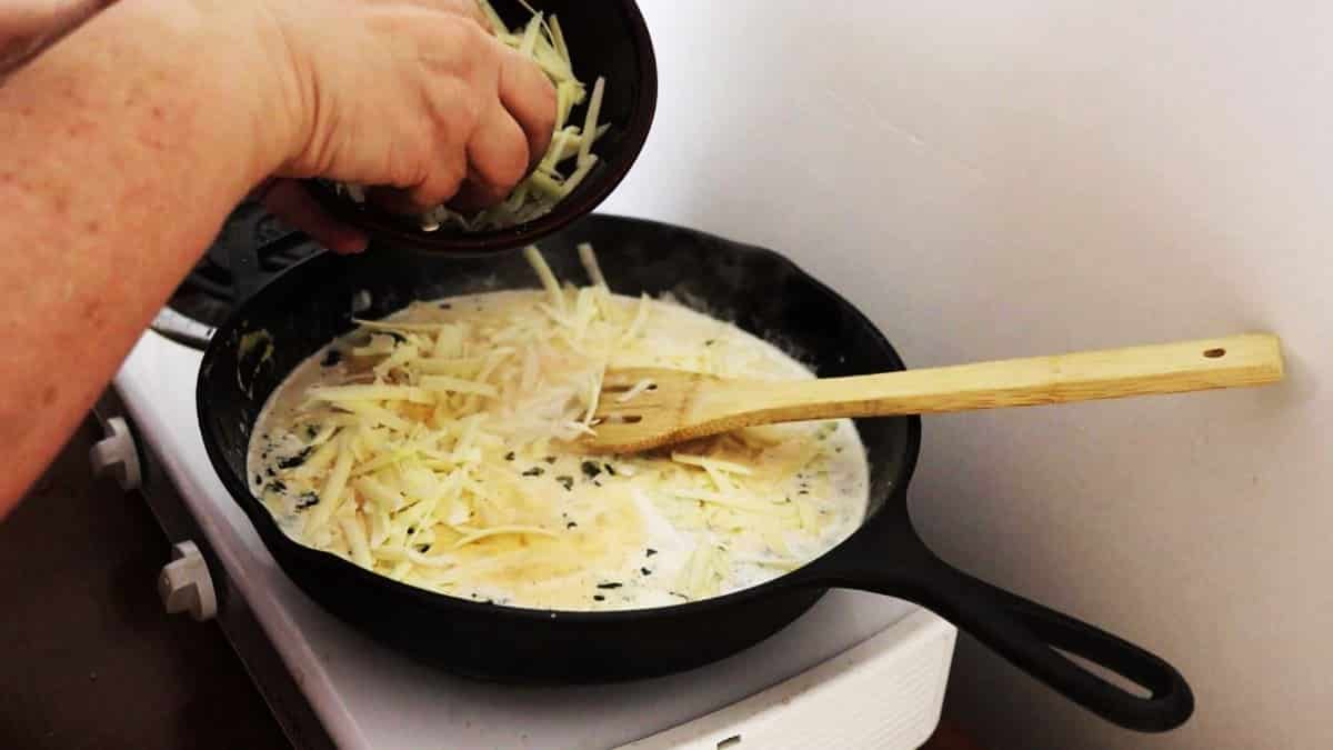 Hand adding cheese from a bowl into a creamy white sauce simmering in a cast iron skillet.