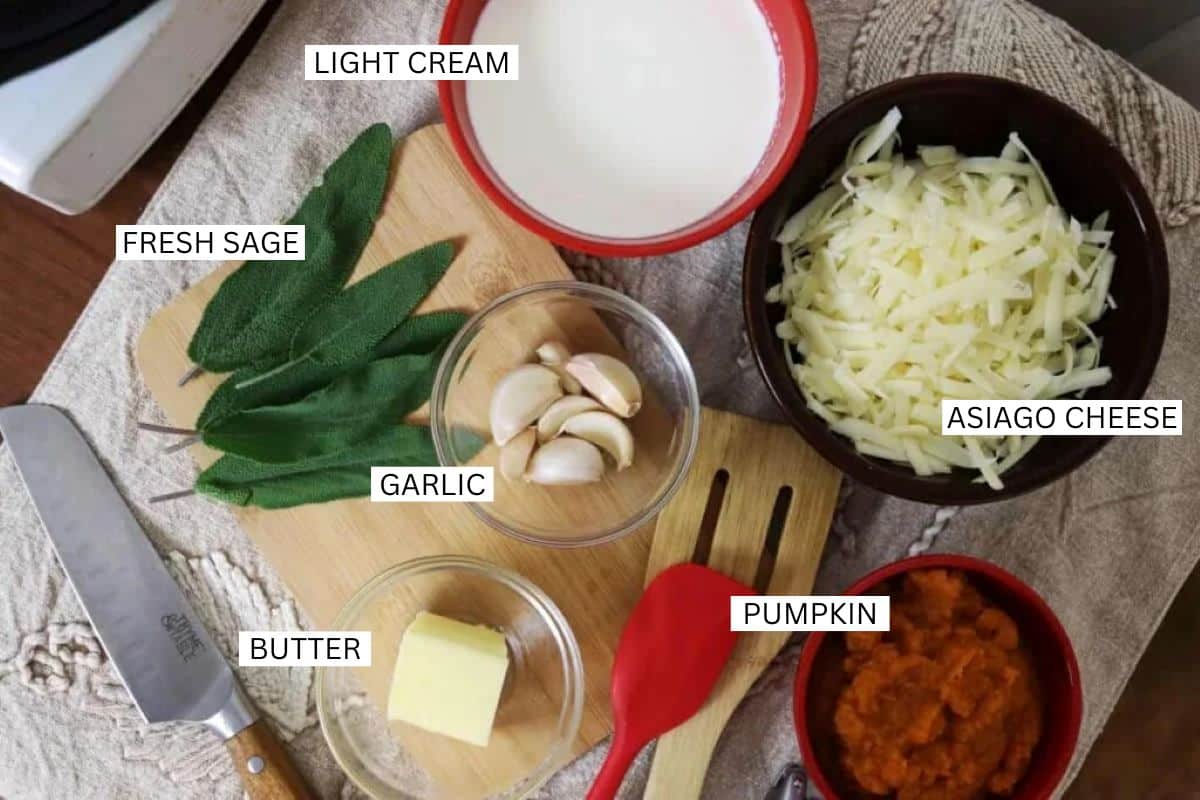 A tabletop displays labeled ingredients: a bowl of light cream, fresh sage leaves, garlic cloves, a bowl of shredded Asiago cheese, a bowl of pumpkin puree, and a pat of butter on a cutting board with a knife and red spatula.
