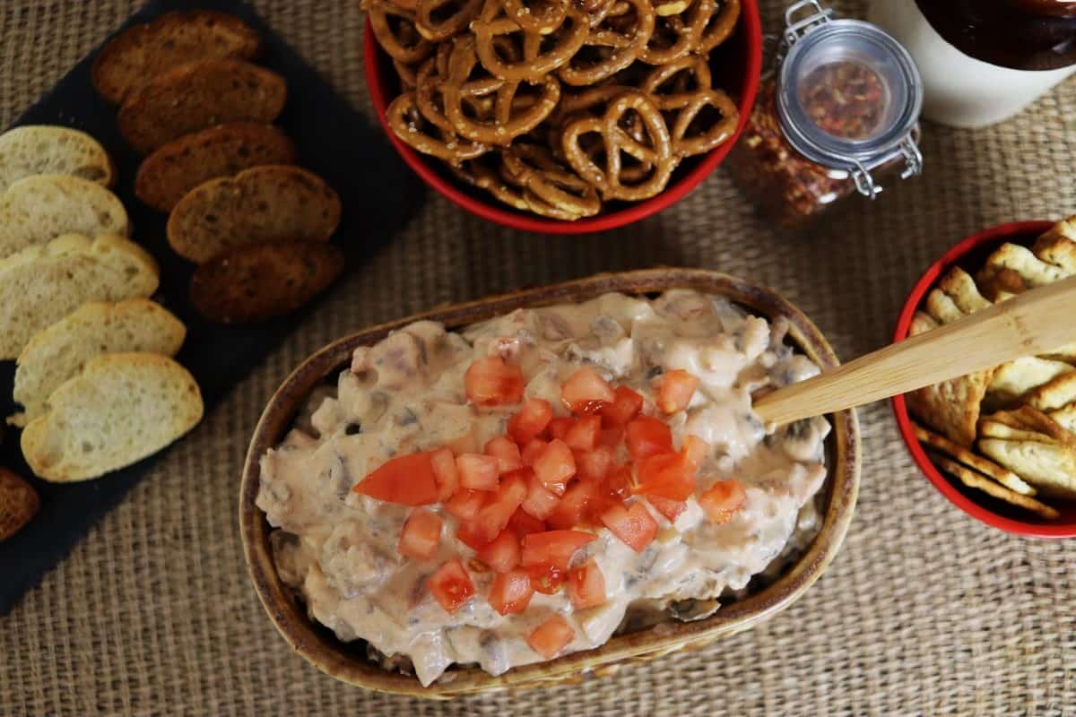 A bowl of creamy dip topped with diced tomatoes, surrounded by pretzels, crackers, toasted bread slices, and a jar of red pepper flakes on a textured surface.