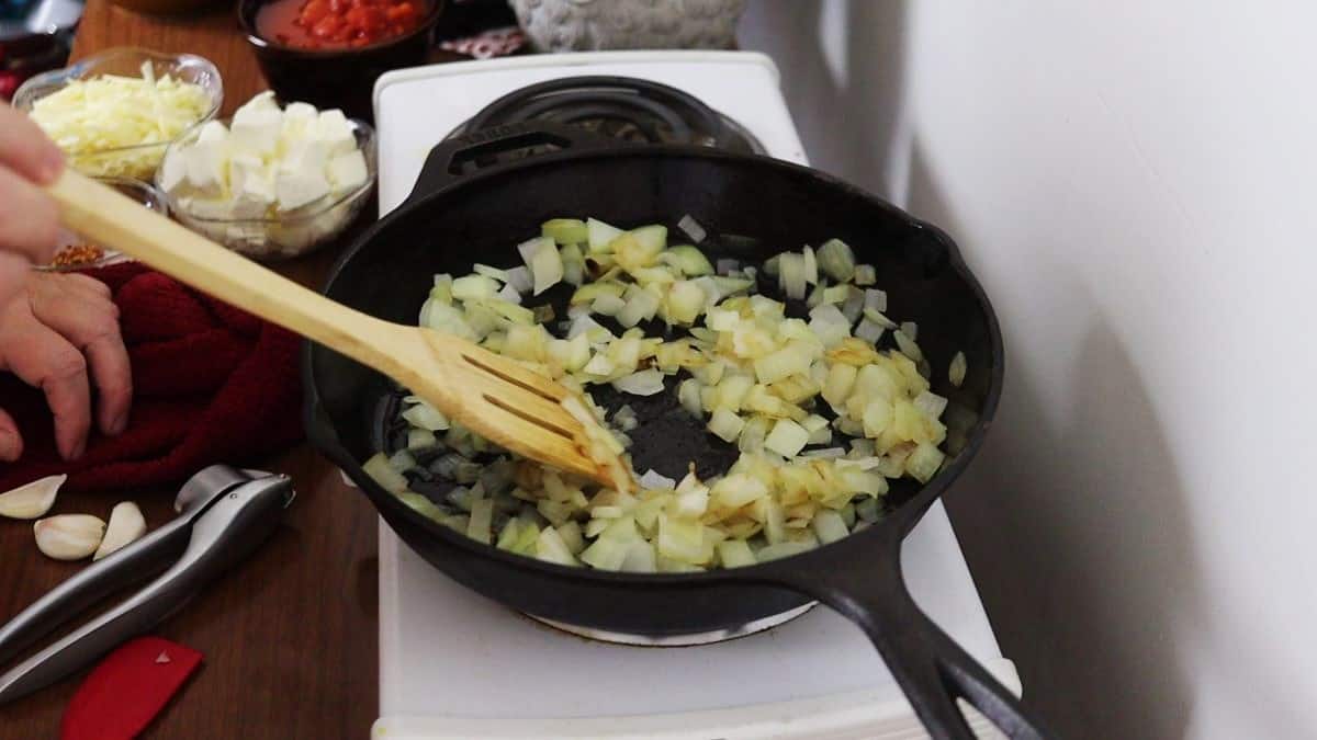 Chopped onions being stirred by a wooden spoon while frying in a cast iron skillet on a stovetop.