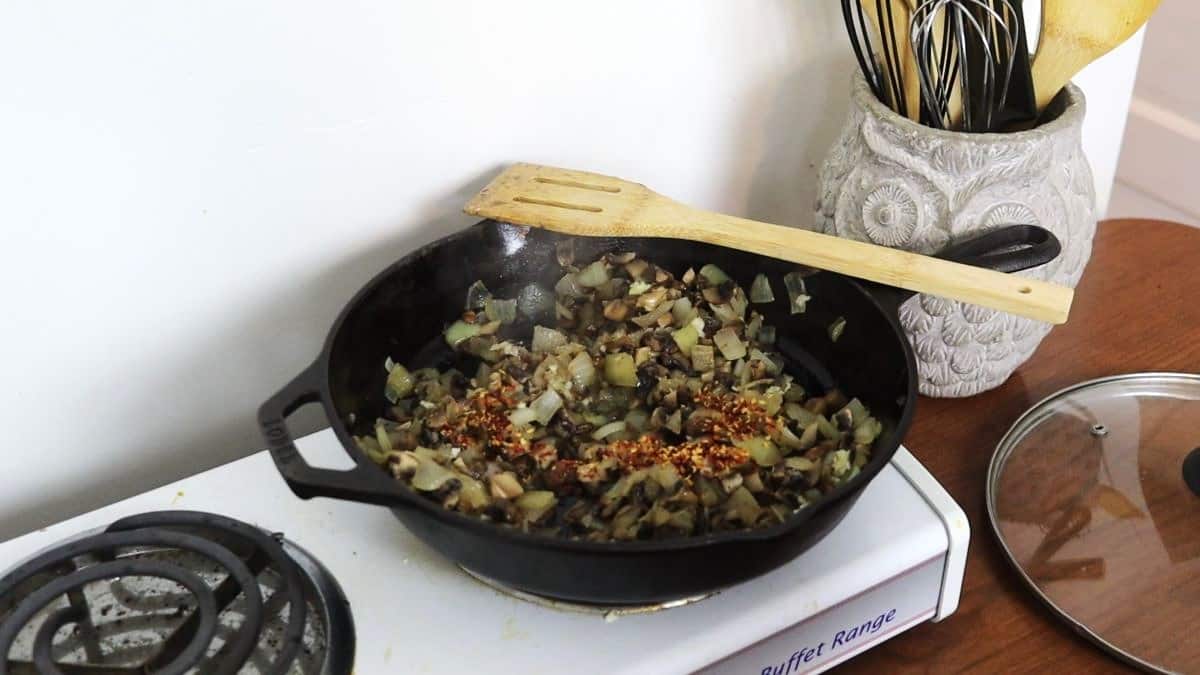 Cooked chopped onions and mushrooms topped with spices cooking in a cast iron skillet on a stovetop with a wooden spoon resting on the edge of the skillet.