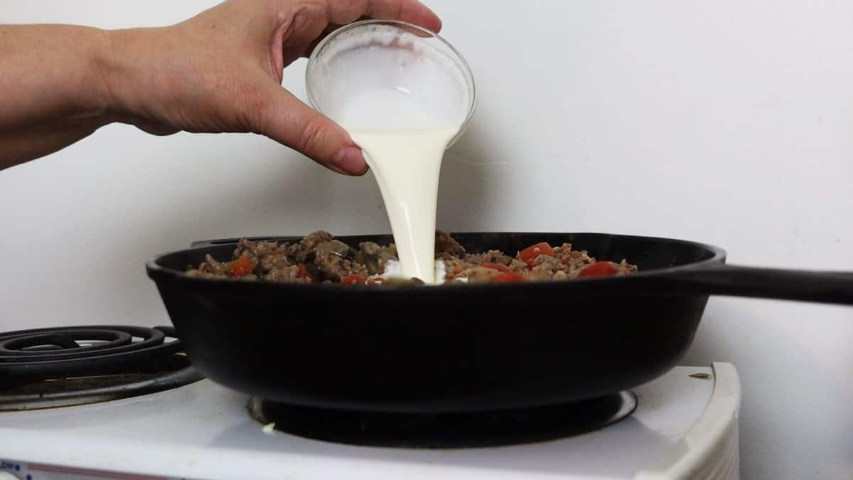 Hand pouring heavy cream into a skillet with sausage and tomatoes in it sitting on a stovetop.