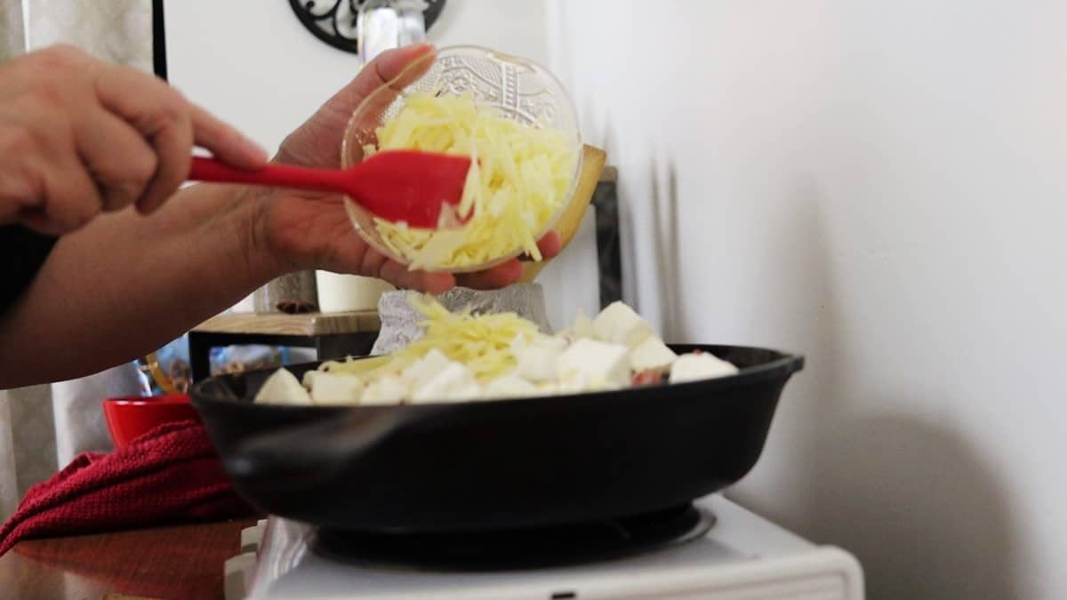 Hands using a red spatula to scrape shredded cheese from a small bowl into a cast iron skillet with chunks of cream cheese in it sitting on a stovetop.