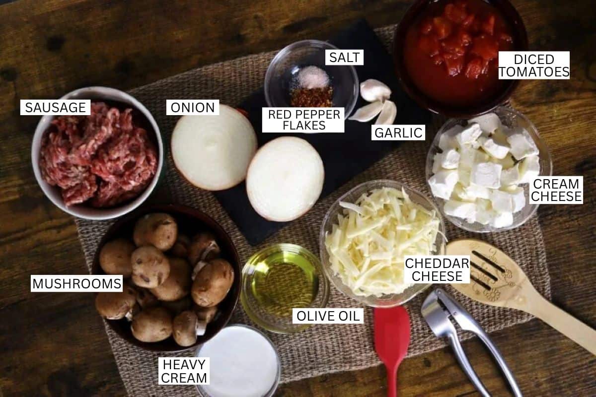 Ingredients on a table: sausage, sliced onion, red pepper flakes, salt, diced tomatoes, garlic, cream cheese, cheddar cheese, mushrooms, olive oil, and heavy cream. Items are labeled with text.