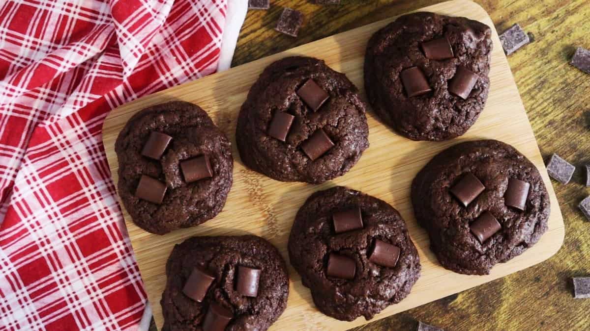 Six double chocolate chip cookies with visible chocolate chunks are arranged on a wooden board next to a red and white plaid cloth, with a few chocolate chunks scattered on the wooden surface.