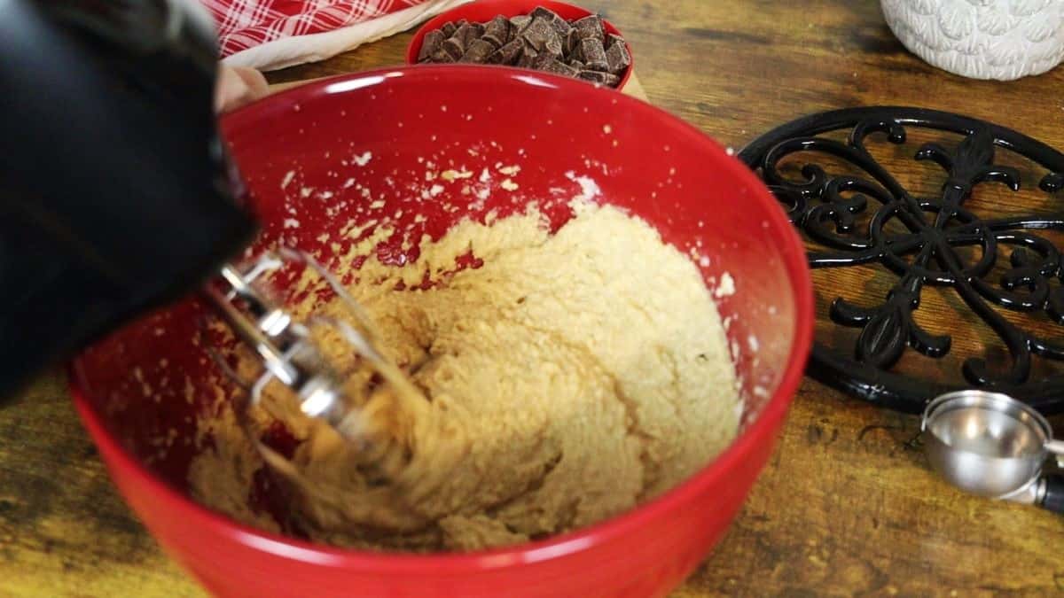 Yellow batter being whipped with beaters in a red bowl with an iron trivet and a dish of chocolate chunks in the background.