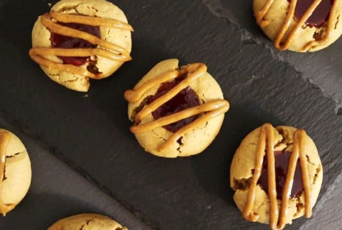 Five peanut butter cookies with jelly centers and a drizzle of peanut butter are arranged on a dark slate surface.