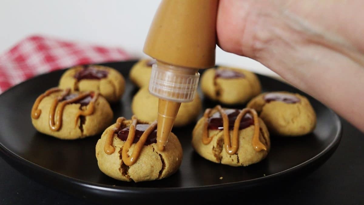 Strips of peanut butter topping being piped from a squeeze bottle over a peanut butter cookie with jelly on the top.